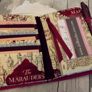 May include: A burgundy fabric notebook with a map of Hogwarts printed on the cover. The notebook has a burgundy ribbon bookmark and a pen holder. The text "The Marauders" is printed on the cover.