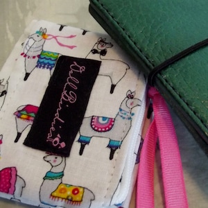 May include: A white fabric wallet with a colorful llama pattern and a black label that reads "Tall Birdiest". Next to it is a green leather journal with a black elastic band and a pink ribbon.