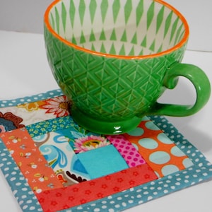 May include: A colourful patchwork fabric coaster with a teal border and a green ceramic mug with a white and green geometric pattern on top.
