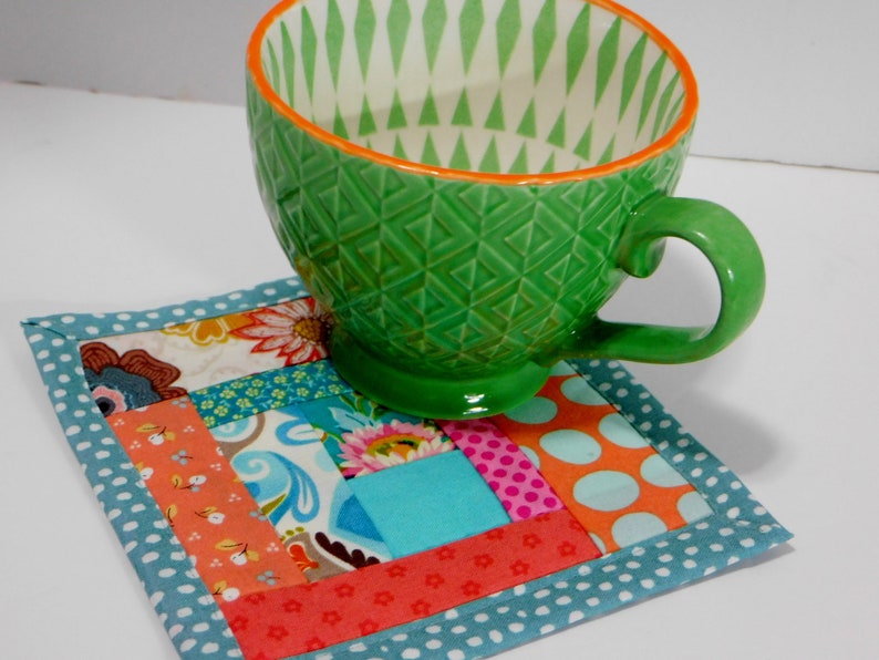 May include: A colourful patchwork fabric coaster with a teal border and a green ceramic mug with a white and green geometric pattern on top.