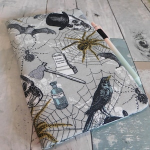 May include: A rectangular fabric notebook cover featuring a light gray background with a repeating pattern of black spiders, spiderwebs, bats, skulls, a tombstone labeled Colden Buried, an axe, and a bottle. A light-colored pen rests on the cover.