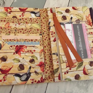 May include: A fabric-covered journal with a zipper closure and a brown strap. The cover features a fall-themed pattern with birds, leaves, and acorns. The journal has a pocket on the inside cover and a business card holder with the text "www.dollbirdies.etsy.com".