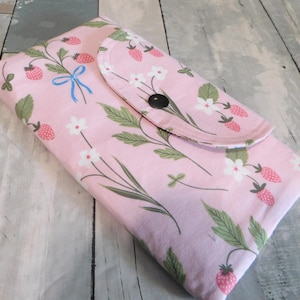 May include: A pink fabric wallet with a flap closure, adorned with a pattern of strawberries, white flowers, and green leaves. A light blue bow accents the design. The wallet is closed with a black button.