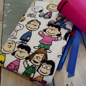 May include: A white pencil case featuring a repeating pattern of Peanuts characters in various poses. A pink cylindrical case, a blue pen, and a blue ribbon are visible. The pencil case is made of fabric.