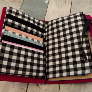 May include: Open magenta-colored journal with black and white checkered fabric lining the inside covers. The left side has card pockets and black fabric stripes. A pen is tucked into the right side. The journal is open, revealing the interior.