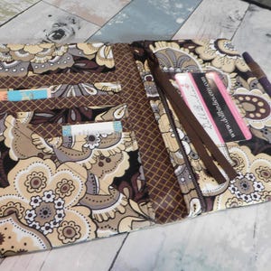 May include: Open floral patterned wallet in shades of beige, brown, and black. The wallet has multiple card slots, a zippered compartment, and a pen holder. A business card is visible inside the wallet.