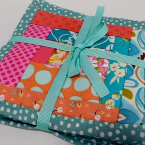 May include: Set of four teal and white polka dot fabric coasters with a patchwork design in shades of pink, orange, and blue. The coasters are tied together with a teal ribbon.