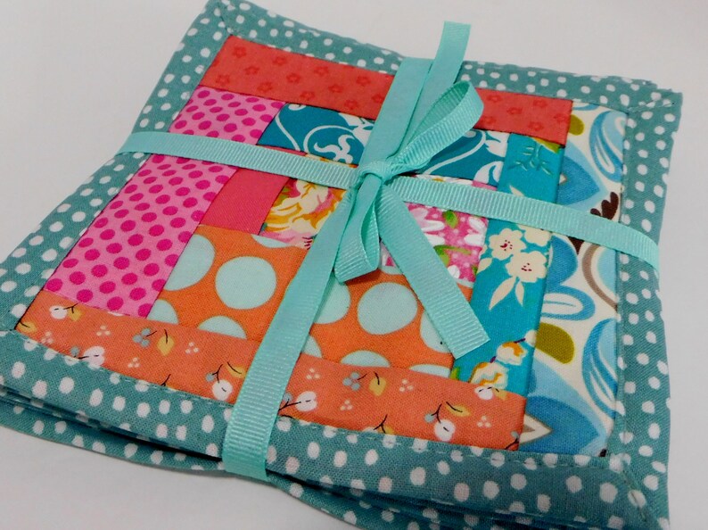 May include: Set of four teal and white polka dot fabric coasters with a patchwork design in shades of pink, orange, and blue. The coasters are tied together with a teal ribbon.