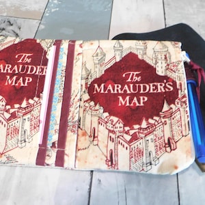 May include: A cream-colored zippered pouch featuring a burgundy design of a castle and the text "The Marauder's Map." The pouch has a burgundy zipper pull and a blue pen. A black notebook is visible in the background.