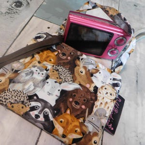 May include: A small fabric pouch featuring a colorful woodland animal print with bears, foxes, owls, and hedgehogs. A bright pink digital camera rests on top of the pouch, which includes a brown fabric strap and a black tag labeled All Birdies.