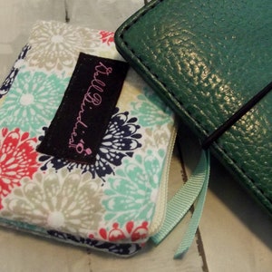 May include: A fabric wallet featuring a floral pattern in red, teal, and navy blue with a black rectangular tag labeled "AllBirdies" and a textured dark green leather-like notebook cover with a black elastic band and a light blue ribbon bookmark.