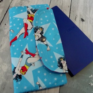 May include: A blue fabric wallet with a Wonder Woman design. The wallet has a flap closure and a blue background with white stars and multiple images of Wonder Woman.