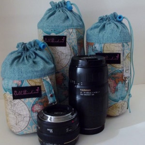 May include: Three cylindrical fabric drawstring pouches featuring a vintage world map print in blue, tan, and cream, each with a black label reading "Bell Birdies". Two black camera lenses are positioned in front of the pouches on a white surface.