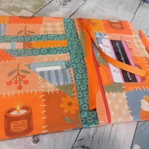 May include: A fabric planner cover featuring a patchwork design with orange, teal, and cream colors. The exterior displays illustrations of candles, berries, and pie slices. It includes multiple interior pockets, a clear card slot, and an orange pen.