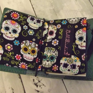 May include: Open green leather journal with a black elastic closure. The interior features a black fabric with a colorful sugar skull pattern. The journal has a black strap attached to the spine. The text "Bell Birdies" is printed on a black label.
