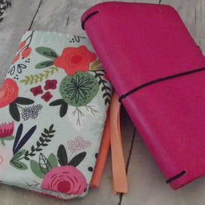 May include: Two journals: a bright pink leather journal with a black elastic closure and a floral fabric journal. The floral journal has a light blue background with red, pink, and green flowers. Both have ribbon bookmarks.