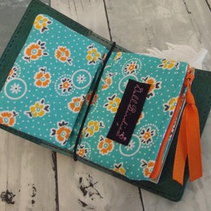 May include: A teal green leather wallet with two pockets lined with turquoise fabric with a white polka dot and orange floral pattern. The wallet has a zipper closure and an orange ribbon.