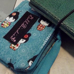 May include: A teal blue fabric wallet with a black label that says "Dull.girl's". The wallet has a zipper closure and a blue strap. The fabric has a pattern of white flowers and Japanese dolls in colorful kimonos.