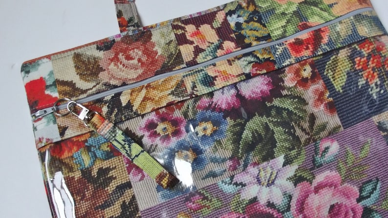 Pode incluir: Uma bolsa tote com padr&atilde;o floral, fecho de correr e uma janela de pl&aacute;stico transparente. A bolsa tem um puxador de z&iacute;per prateado com um chaveiro e uma al&ccedil;a verde e amarela.
