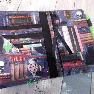 May include: Open book cover with a spooky library print featuring skulls, potion bottles, and a black cat. The cover has a black ribbon closure and a green pen is attached to the side. The text "Vampire Blood" is visible.