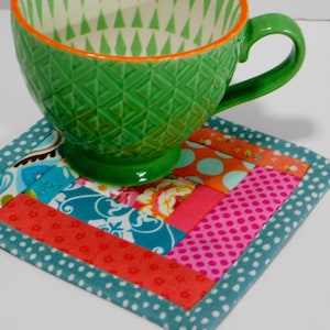 May include: A colourful patchwork fabric coaster with a teal border and white polka dots. A green ceramic mug with a geometric pattern sits on top of the coaster.