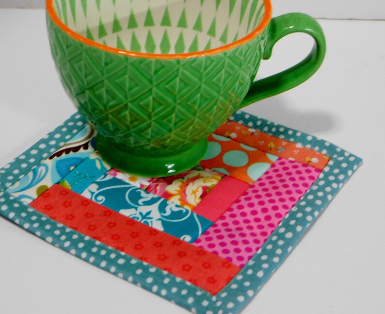 May include: A colourful patchwork fabric coaster with a teal border and white polka dots. A green ceramic mug with a geometric pattern sits on top of the coaster.