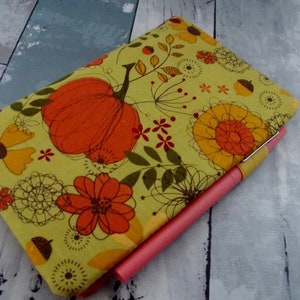 May include: A notebook with a fall-themed design featuring an orange pumpkin, yellow and orange flowers, and brown acorns on a light green background. A pink pen rests on the notebook.