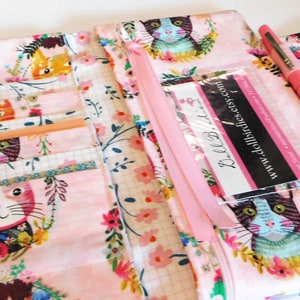 May include: A pink fabric pen case with a cat print. The case is open, revealing pockets and a pencil. A pink pen rests on the case. The cat design features colorful floral wreaths. The case has a pink ribbon and a clear pocket with a business card.