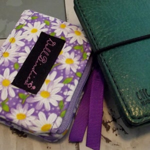 May include: Two small wallets, one with a purple daisy print and a black label that says "It's.a.girl." and a purple ribbon, and the other a dark green leather wallet with the text "Chic Sparrow Made in the USA."