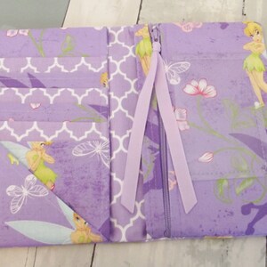 May include: Open, quilted wallet in shades of purple, featuring a fairy and floral pattern. The wallet has multiple card slots, a zippered compartment, and a purple pen. The design includes a white quatrefoil pattern and a light purple ribbon.