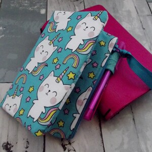 May include: A teal pencil case with a zipper, featuring a repeating pattern of white unicorn cats, rainbows, and stars. A pink pen is visible inside. A pink notebook is in the background.