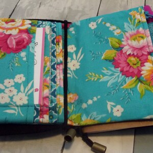 May include: Open journal with a black cover and a floral pattern on the inside. The floral pattern is a teal background with pink and white flowers. The journal has card slots and a pen holder with a teal ribbon.