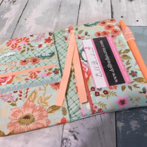 May include: Open floral fabric journal with peach-colored accents. The journal features a floral pattern in shades of peach, pink, and green. A peach ribbon and an orange pen are included. The journal has a business card with the text "Doll Birdies" and a website address.