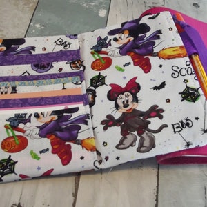 May include: A white fabric zippered pouch with a Halloween theme. The pouch features a repeating pattern of Mickey Mouse and Minnie Mouse dressed as witches, bats, spiders, and the words "Boo" and "Scare". The pouch has multiple pockets and a purple ribbon.