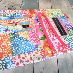 May include: A colorful, patchwork fabric wallet with a floral pattern in shades of blue, orange, pink, and green. The wallet has multiple card slots and a bright pink ribbon closure. A business card with the website address is visible.