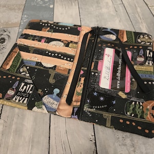 May include: A fabric planner cover featuring a black background with a colorful pattern of potion bottles, skulls, and pumpkins. The interior includes multiple card slots, a clear ID window with a Doll Birdies card, and a pen loop holding a light pen.
