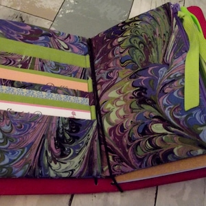 May include: A purple and green marbled fabric notebook with a zippered pocket and a green ribbon. The notebook has several pockets for holding cards or notes.