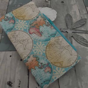 May include: A rectangular journal or notebook with a world map design. The cover features a light blue background with circular map illustrations in shades of beige, orange, and blue. The notebook is closed, with a turquoise edge visible.