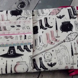 May include: A white fabric pouch with a black and pink witch-themed pattern. The pattern includes witch boots, hats, cauldrons, cats, and candles. The pouch has multiple pockets and is tied with a pink ribbon.