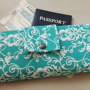 May include: A teal travel wallet with a white floral pattern. The wallet has a flap with a snap closure and a pocket for a passport. The text "PASSPORT" is visible on the passport.