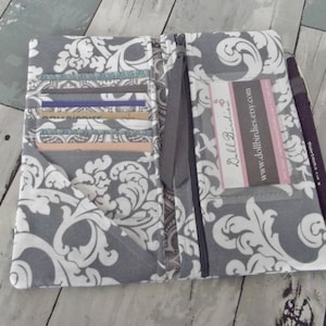 May include: A gray and white patterned fabric wallet with a zipper closure. The wallet has multiple card slots and a clear plastic window for an ID card. The wallet also has a small pocket for cash or receipts. The wallet is open and the inside is visible. The text "Dollbirdies" and "www.dollbirdies.etsy.com" is printed on the inside of the wallet.