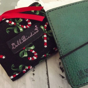 May include: A black zippered pouch with a candy cane and bow pattern, a red ribbon, and a green leather journal with a black elastic closure. The pouch has the text "Doll Birdies" on a black label. The journal has the text "Chic Sparrow Made in the U.S.A."