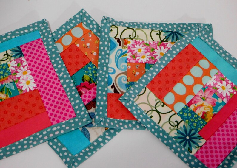 May include: Four handmade fabric coasters with a patchwork design. The coasters are made with a variety of colourful fabrics, including pink, orange, blue, and white. The coasters are edged with a teal fabric with white polka dots.