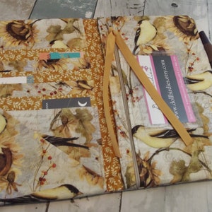 May include: Open fabric journal with a sunflower and bird print. The journal has multiple pockets, a zipper closure, and a ribbon bookmark. A brown pen rests on the side. The business card reads: www.dollbirdies.etsy.com.