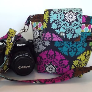 May include: A black camera with a Canon lens is attached to a colorful fabric camera bag with a black strap. The bag has a floral pattern with pink, blue, green, and yellow accents.