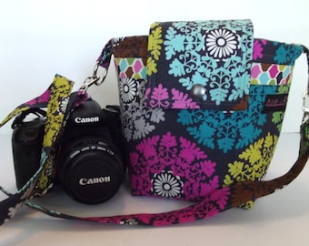 May include: A black camera with a Canon lens is attached to a colorful fabric camera bag with a black strap. The bag has a floral pattern with pink, blue, green, and yellow accents.
