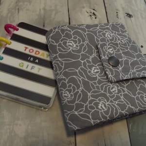 May include: A gray fabric notebook cover with a white floral pattern and a button closure. The cover is closed and a black and white striped notebook with the text "TODAY IS A GIFT" is visible inside.