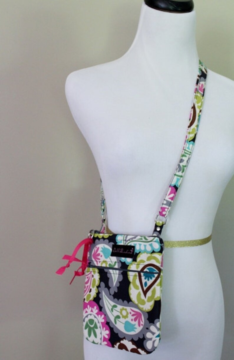 K&ouml;nnte beinhalten: Eine kleine Umh&auml;ngetasche mit Paisley-Muster in Schwarz und Wei&szlig;, einem rosa Schleifen-Detail und einem silbernen Kettenriemen. Die Tasche hat einen Rei&szlig;verschluss und ein kleines Fach auf der Vorderseite. Die Tasche ist von der Marke "Bulle & Co."
