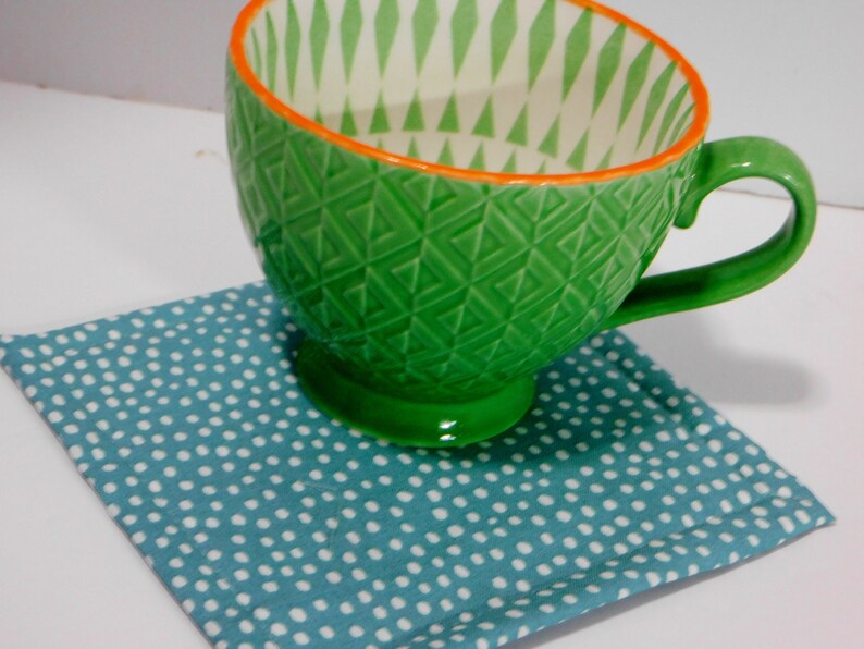 May include: A green ceramic mug with a white and green geometric pattern and an orange rim. The mug is sitting on a blue and white polka dot coaster.