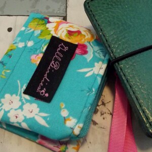 May include: A turquoise fabric wallet with a floral pattern and a black label that says "Lilly Princess". The wallet is next to a teal leather wallet with a pink ribbon.